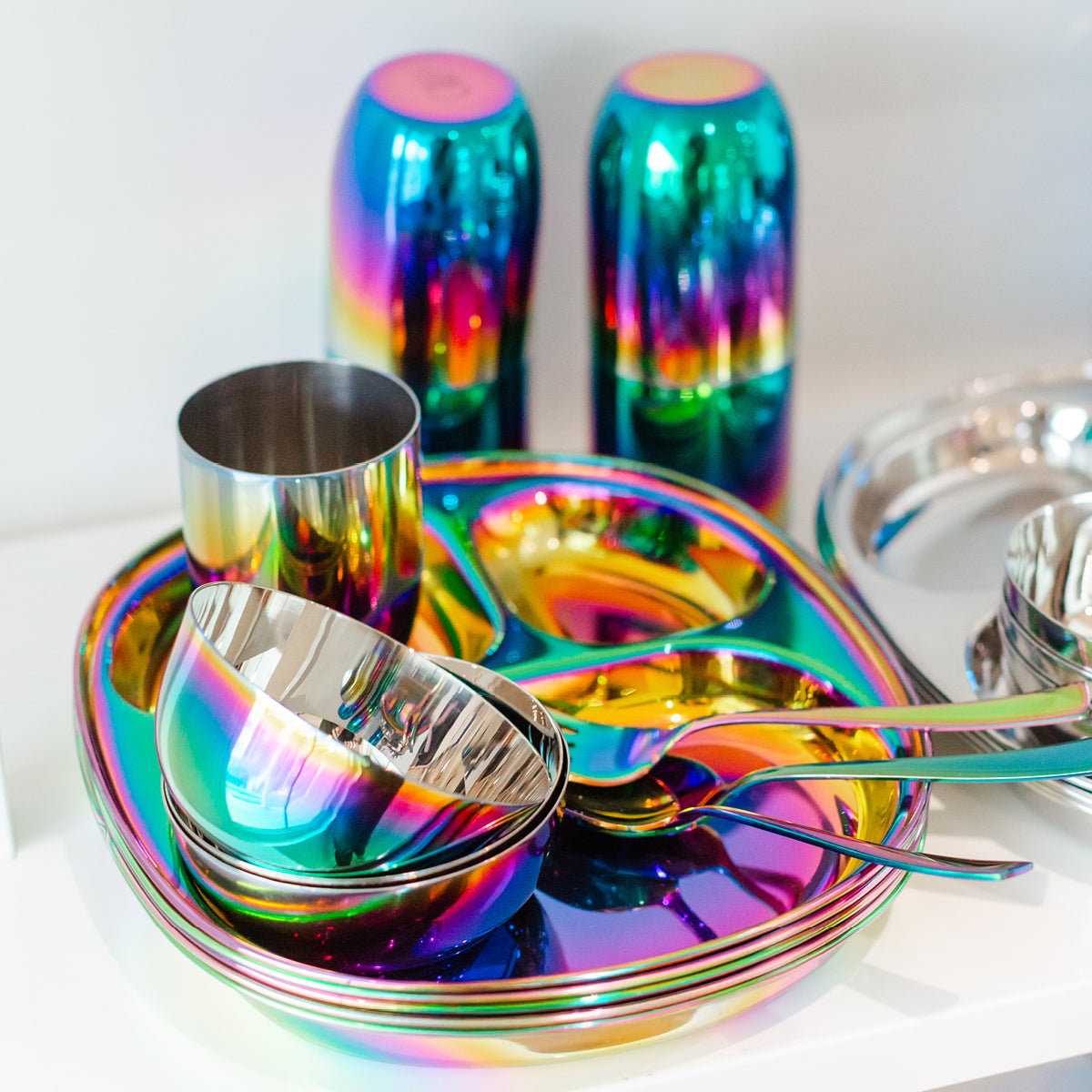 Set of colorful stainless steel dinnerware including plates, bowls, cups, forks, and spoons on a white background.