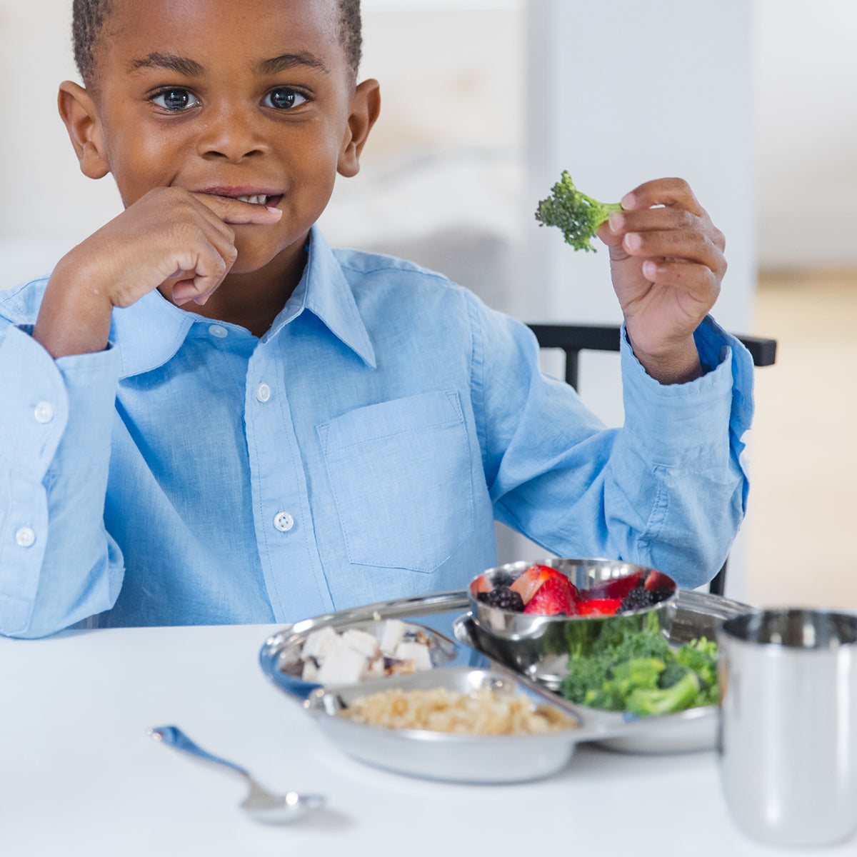 Child in a blue shirt holding a piece of broccoli at a table with a healthy meal on stainless steel divided plate, 8 ounce bowl, fork, spoon, and 8 ounce cup.
