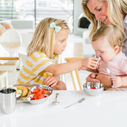 Stainless steel divided plate, 4 ounce training cup, bowl, and infant spoons. Little girl helping her baby sister drink from training cup.