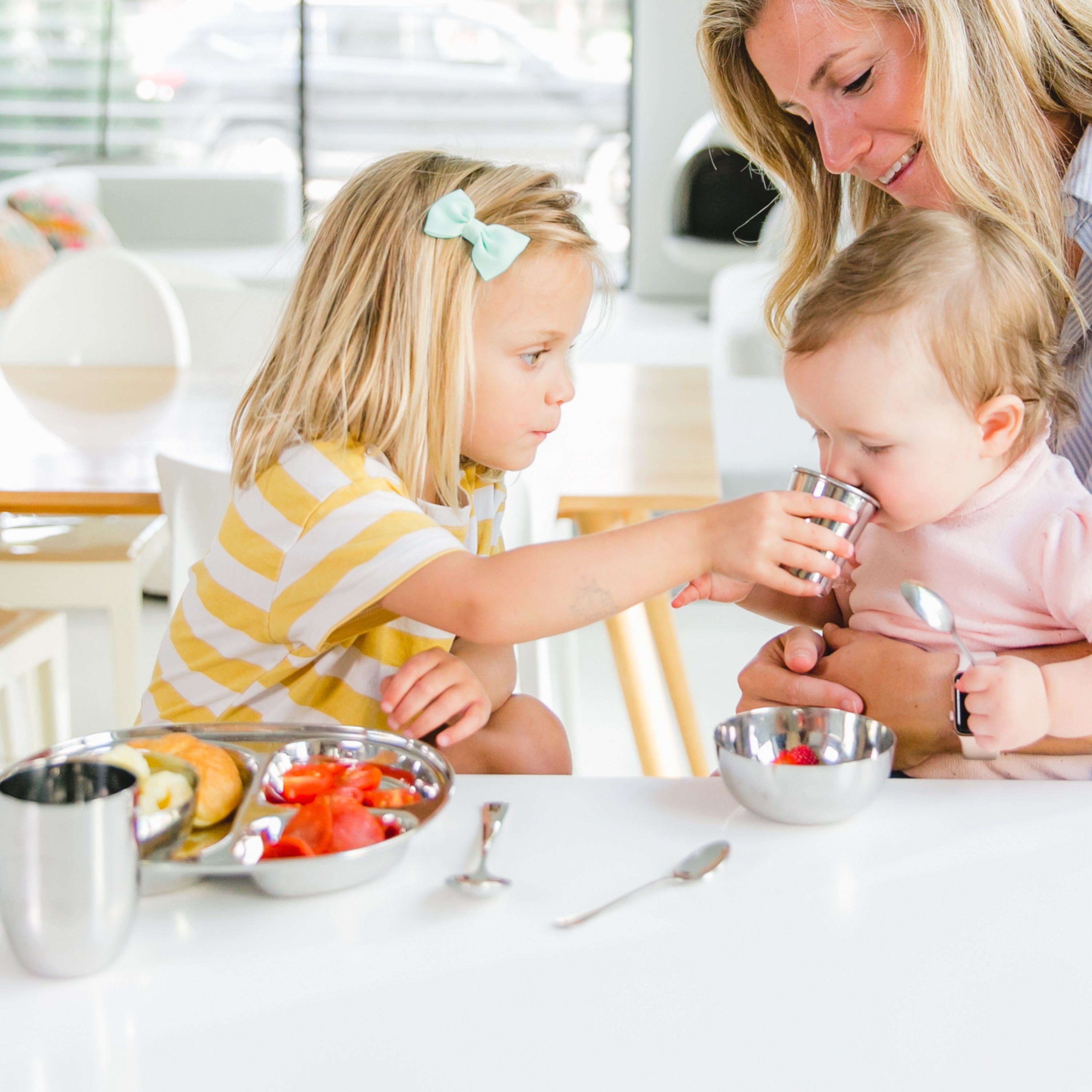 Stainless steel divided plate, 4 ounce training cup, bowl, and infant spoons. Little girl helping her baby sister drink from training cup.
