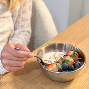 Person eating yogurt with berries and nuts in a stainless steel 12 ounce bowl on a wooden table.