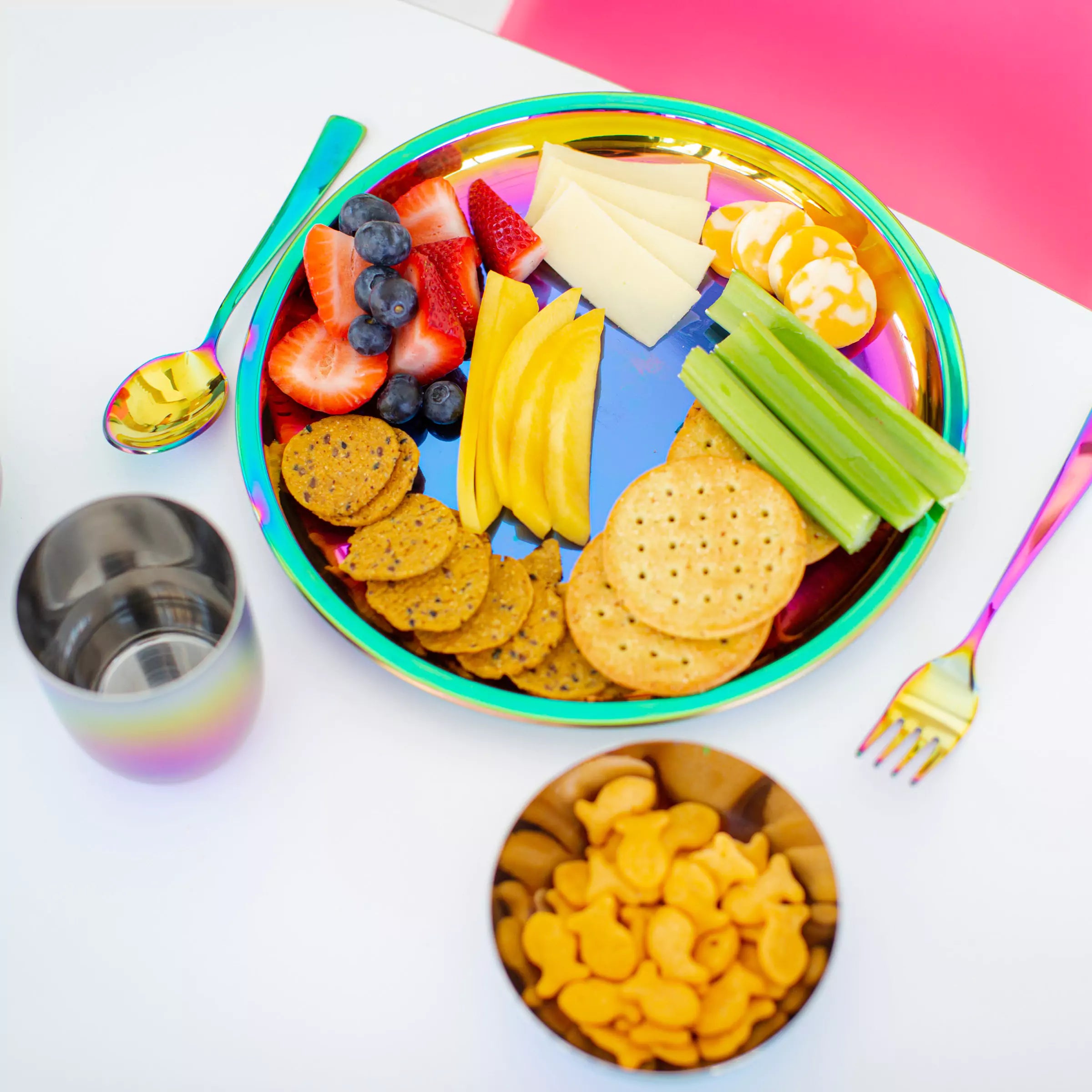 Rainbow Ahimsa stainless steel 9" Purposeful Plate with food (strawberries, blueberries, celery, cheese, crackers) with 8 ounce stainless steel Rainbow Conscious Cup and 8 ounce round bowl with goldfish crackers, and Rainbow Utensil Set (fork and spoon)