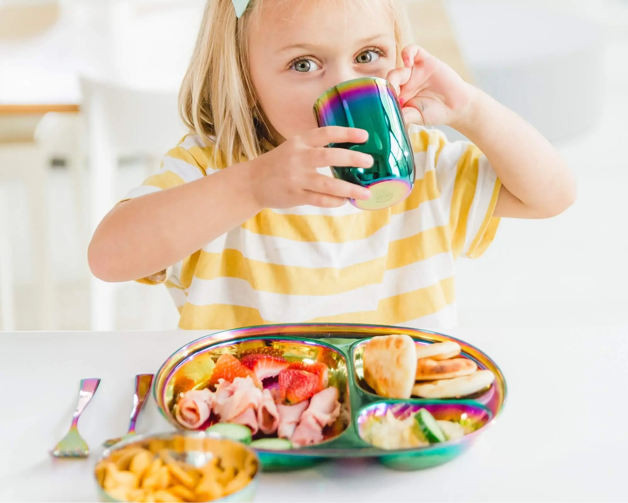Child drinking from a colorful stainless steel tumbler with a healthy dinner in a divided plate for toddlers.