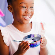 Little girl with a big smile eating yogurt and blackberries from an 8 ounce Iridescent-Blue round bowl and spoon.