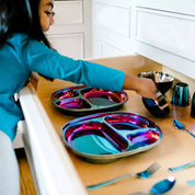Kids plates & bowls set. Little girl in a kitchen, opening a drawer of dishes - the dishes are Ahimsa's Iridescent-Blue stainless steel dishes including: Balanced Bites plates, Smart Snacking Bowls (12 ounces), Starter Sips Cups (4 ounces), round bowls (8 ounces), forks, and spoons. All Ahimsa dishes are dishwasher safe.