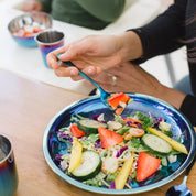 Adult woman eating a salad on a blue 9 inch round Purposeful Plate, She is next to a baby in a high chair with a 4 ounce Starter Sips Cup and an 8 ounce round bowl.