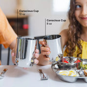 Two children holding stainless steel cups in Classic color, labeled 'Conscious Cup' with sizes 12 oz and 8 oz, on a table with food.
