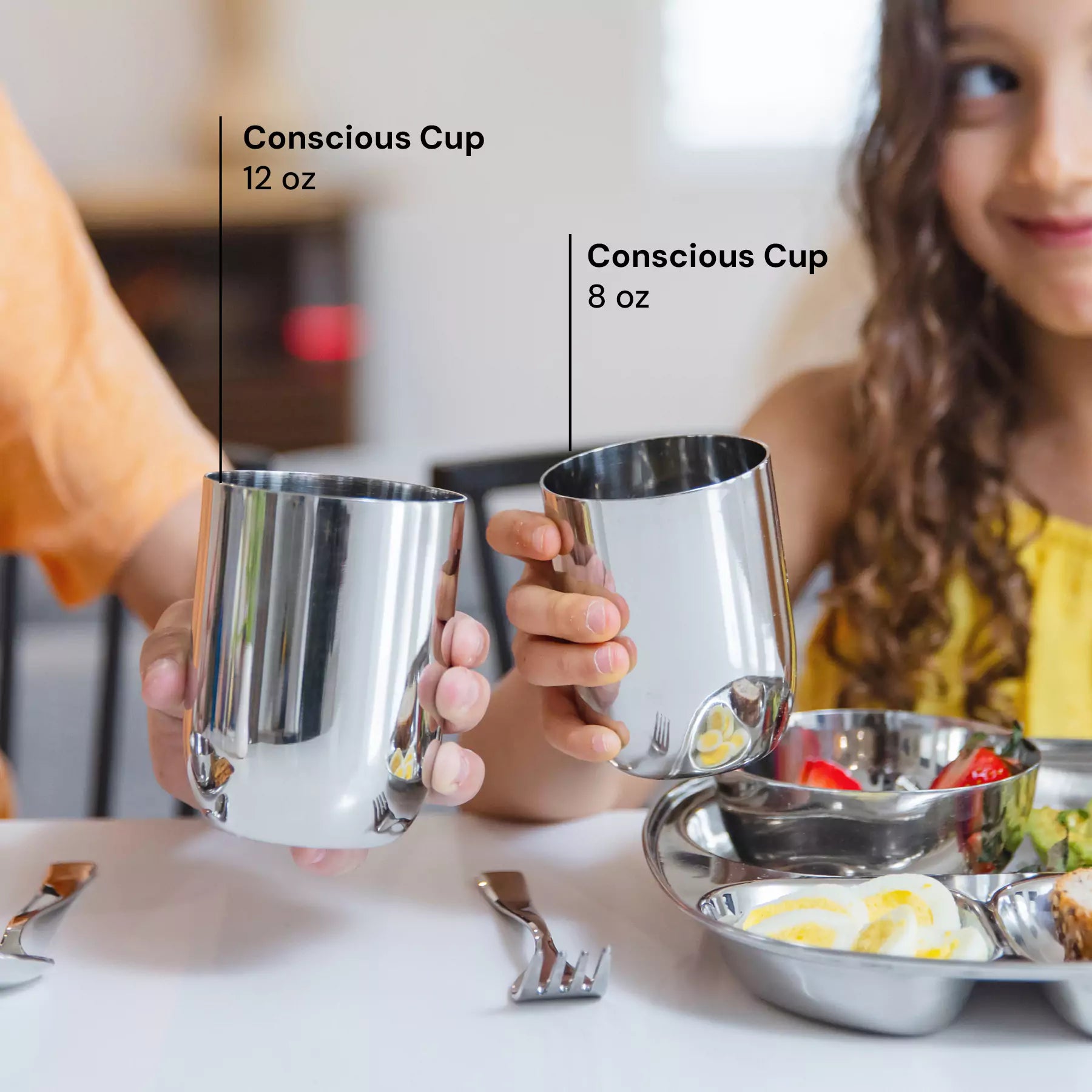 Two children holding stainless steel cups in Classic color, labeled 'Conscious Cup' with sizes 12 oz and 8 oz, on a table with food.