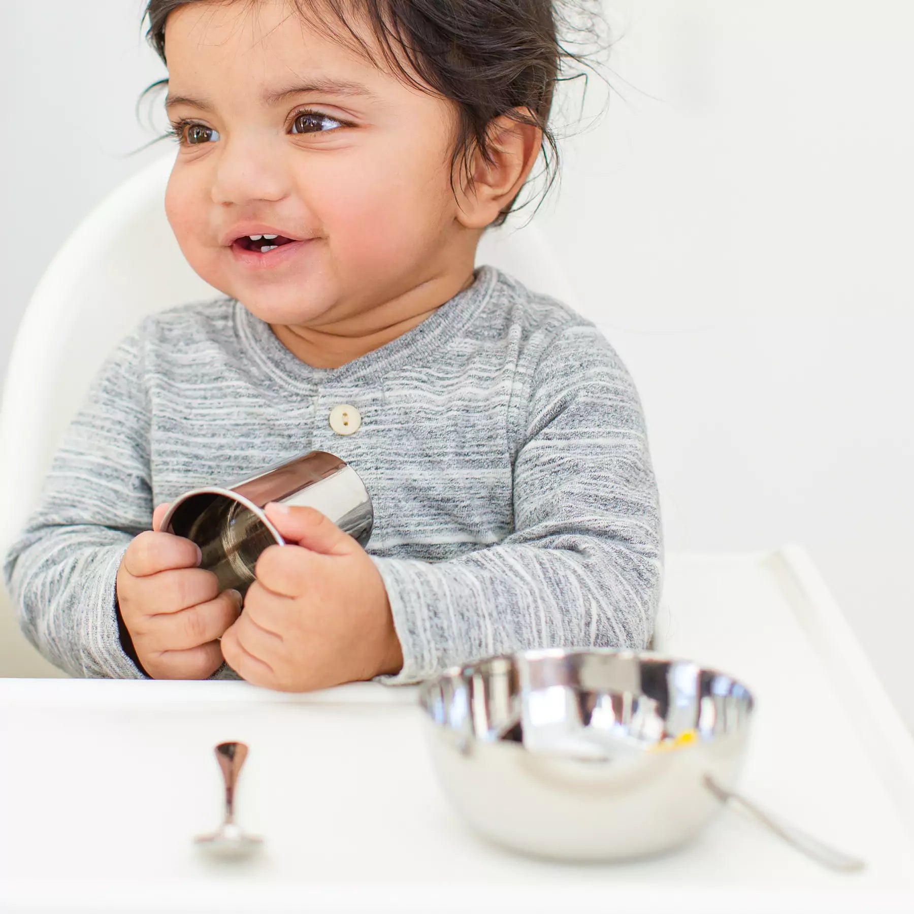 Child holding a metal cup with a stainless steel bowl and spoon on his high chair tray