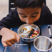 Child eating from a metal bowl with berries, granola, and yogurt, sitting at a table with Ahimsa's Essentials 16 ounce bowl, spoon, and stainless steel cup.