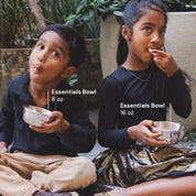 Two children sitting outdoors, each holding a stainless steel bowl labeled to show sizing of 8 ounce bowl and 16 ounce bowl