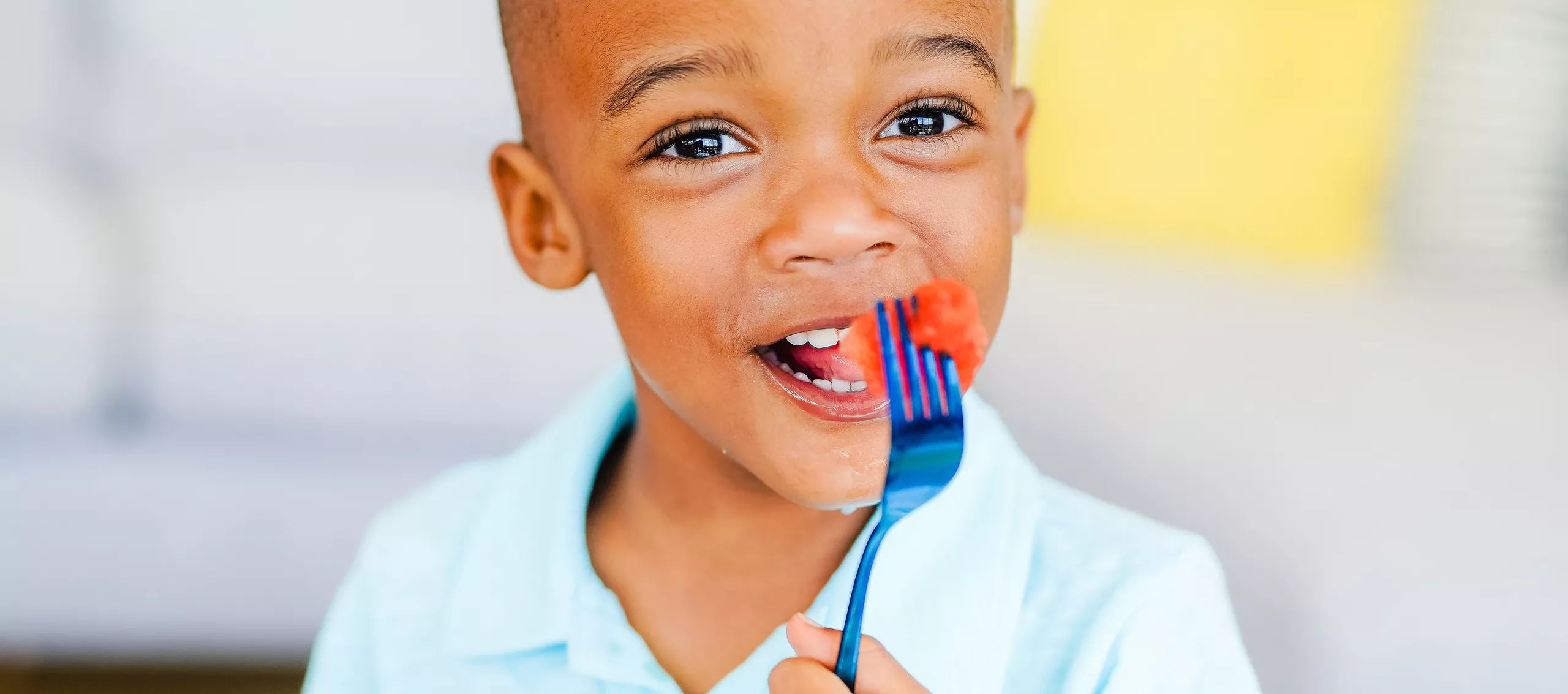 A young boy wearing a light blue polo shirt, holding a blue stainless steel fork with a piece of watermelon on it near his mouth, set against a soft-focus indoor background.