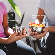 Two teenagers holding stainless steel bowls labeled 'Essentials Bowl 8 oz' and 'Essentials Bowl 16 oz' filled with colorful food to show the sizing of these beautiful, durable, dishwasher safe bowls. 