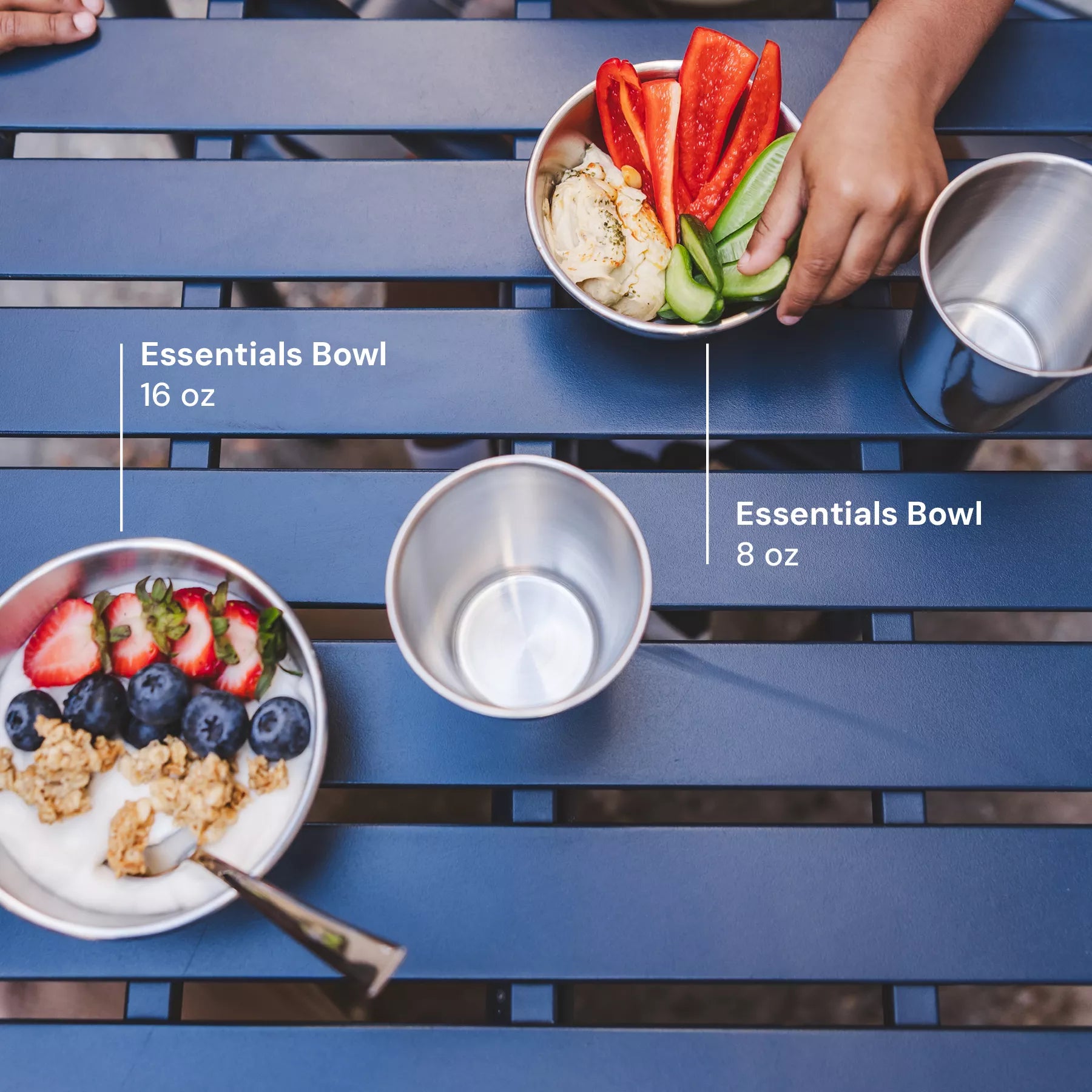 Two bowls labeled 'Essentials Bowl 16 oz' and 'Essentials Bowl 8 oz' on a blue picnic table with food. Shows size of the bowls. 