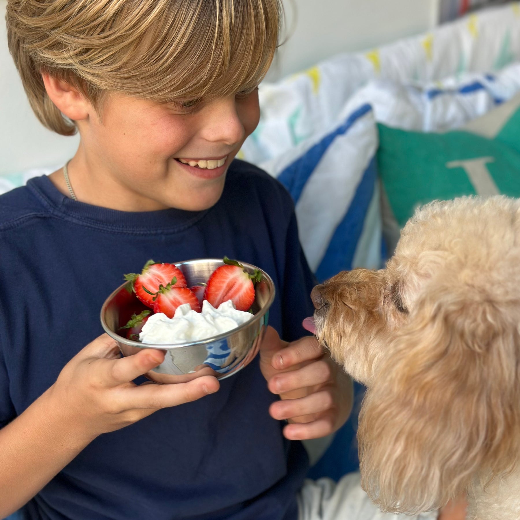 Young boy holding a stainless steel bowl of strawberries and cream and a dog looking at it. 