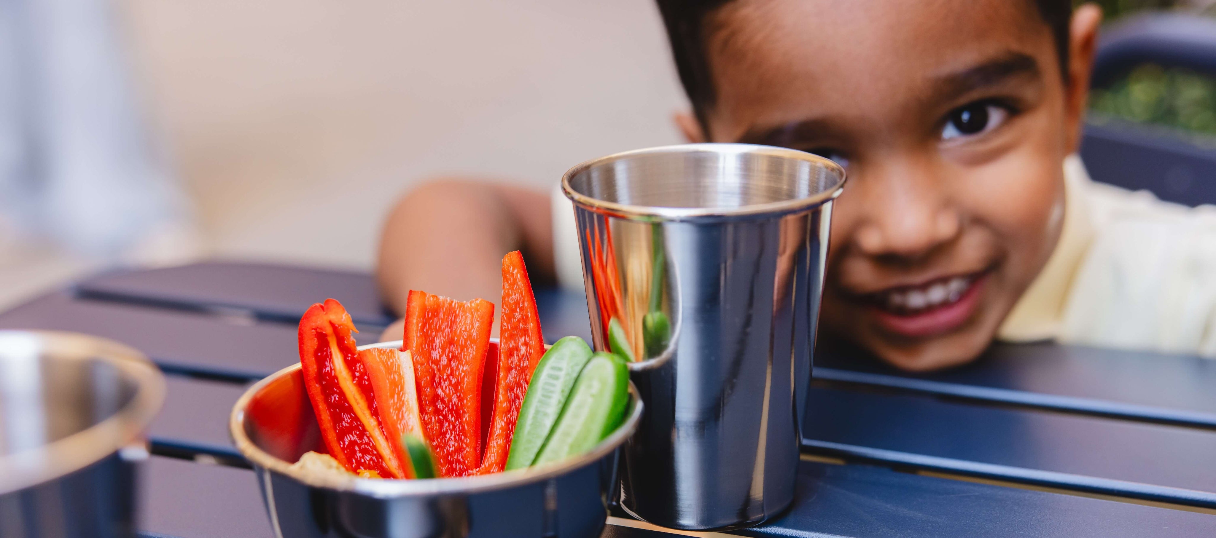 Child at a table with a small stainless steel bowl of vegetables and a stainless steel metal drinking cup, outdoor setting
