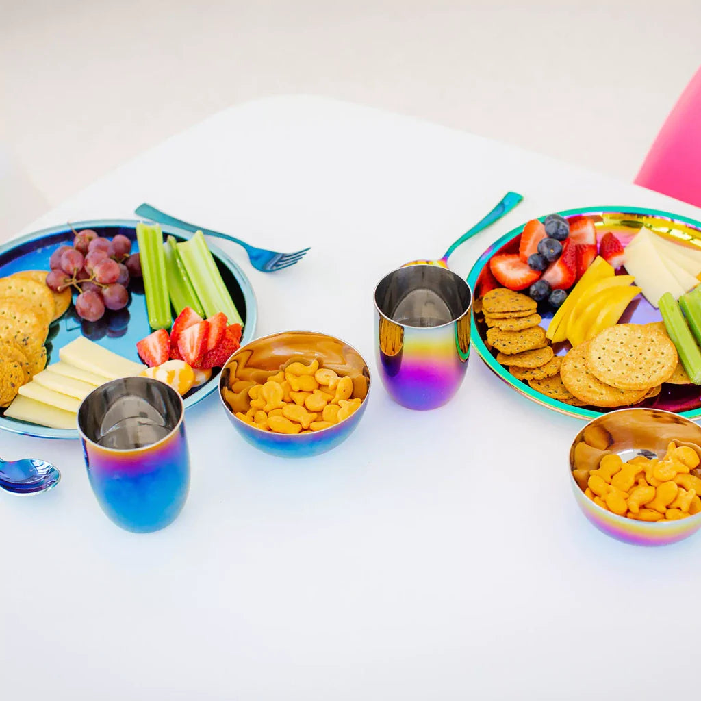 2 place settings on a table - the left side is Blue and the right side is Rainbow. Each place setting includes a 9 inch round Purposeful Plate (with crackers, fruit, cheese, and celery), an 8 ounce Conscious Cup with water, fork, spoon, and an 8 ounce round bowl with goldfish crackers