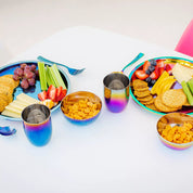 2 place settings on a table - the left side is Blue and the right side is Rainbow. Each place setting includes a 9 inch round Purposeful Plate (with crackers, fruit, cheese, and celery), an 8 ounce Conscious Cup with water, fork, spoon, and an 8 ounce round bowl with goldfish crackers