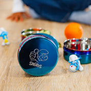 Blue stainless steel food container with 'Smurfs' logo on a wooden surface with Smurf figurines.