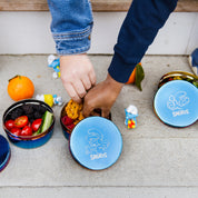 Children's hands reaching into a Smurfs-themed lunch stainless steel containers with pretzels, tomatoes, blackberries, raspberries, and cucumbers.