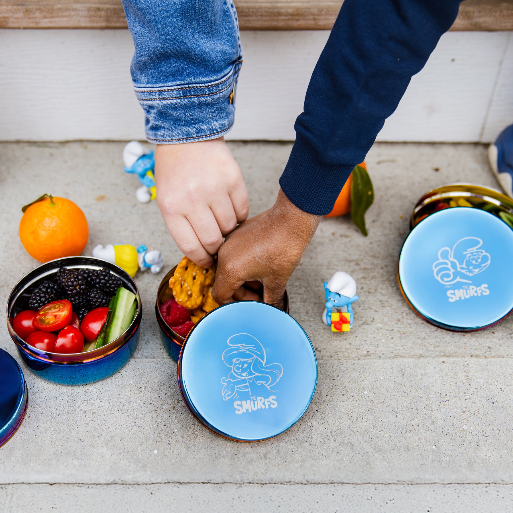 Children's hands reaching into a Smurfs-themed lunch stainless steel containers with pretzels, tomatoes, blackberries, raspberries, and cucumbers.