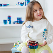 Young girl wearing a 'The Smurfs' sweatshirt, holding a blue iridescent, Ahimsa Large Circle stainless steel food container with berries, in a room with shelves displaying more dishes.