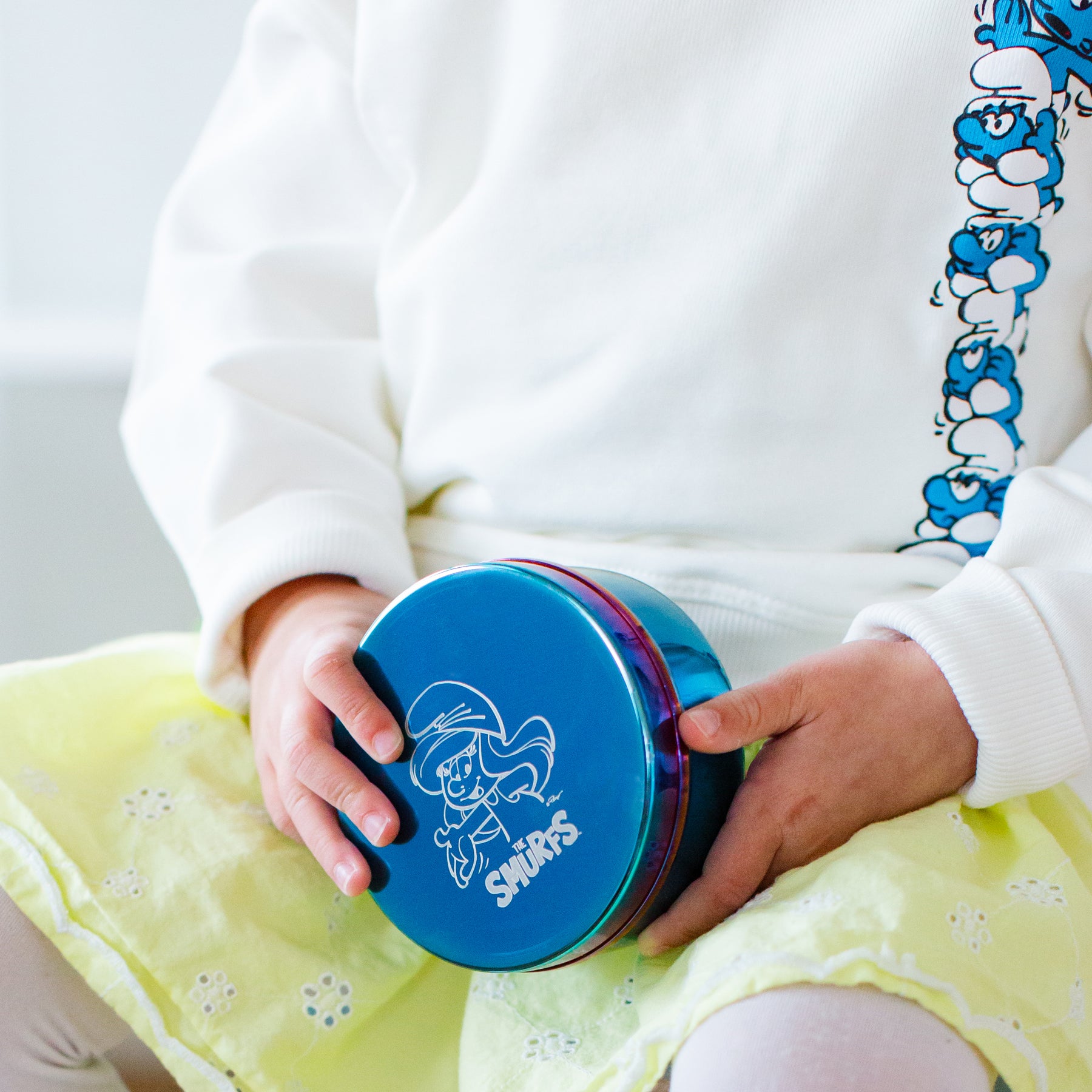Child holding a blue stainless steel food container with a Smurfs logo and Smurfette, wearing a white sweater with Smurf design.