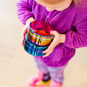 Child holding a stack of colorful stainless steel food containers in rainbow and blue against a neutral background