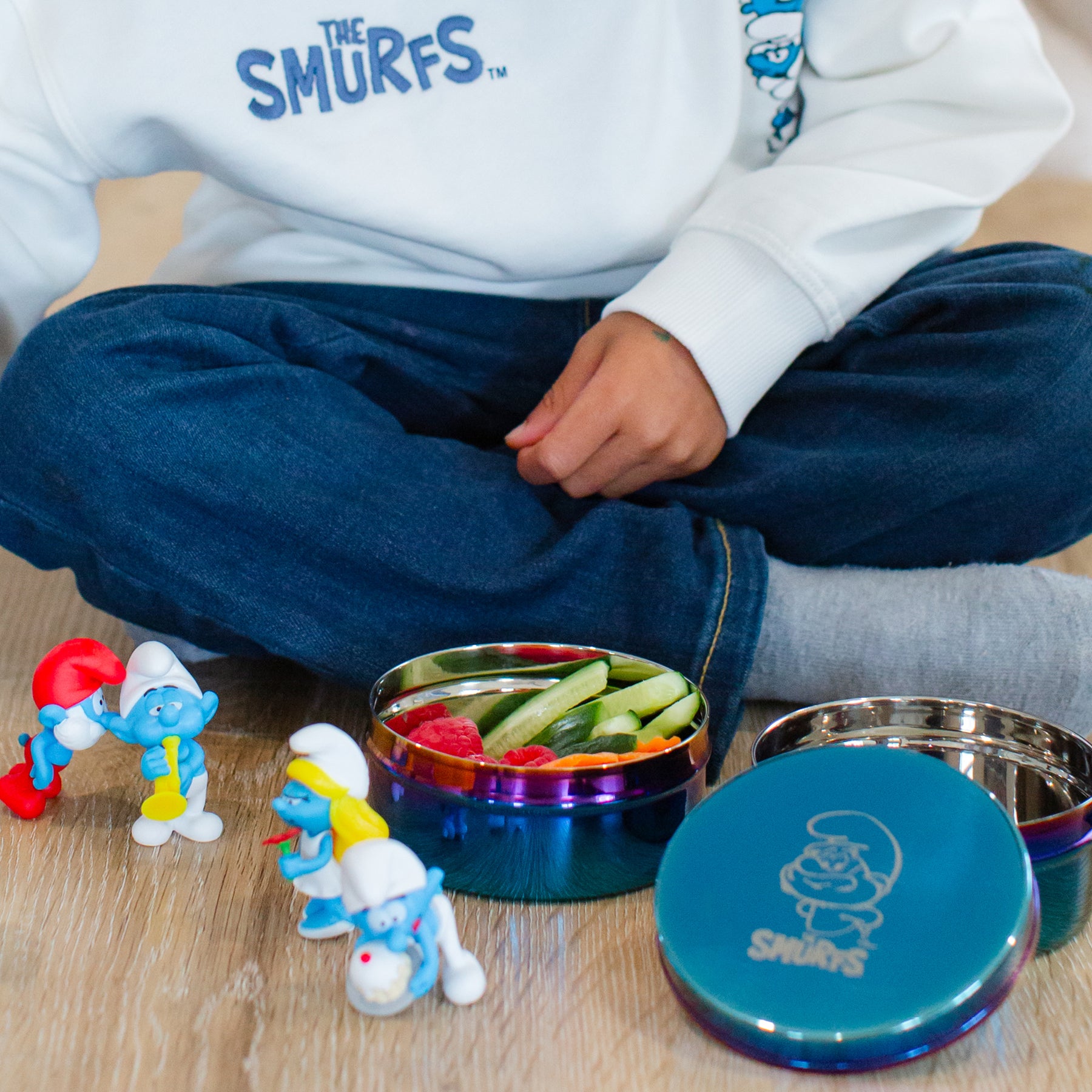 Little boy wearing a Smurfs hoodie with Smurfs toys and stainless steel food containers with raspberries, cucumbers, and carrots