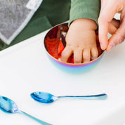 toddler self feeding from a baby bowl with 2 blue baby spoons on the table