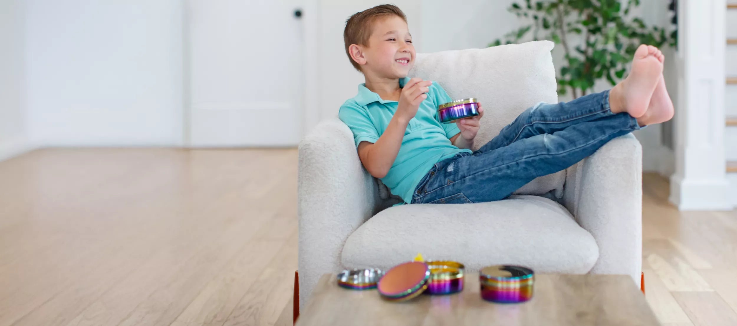 A young boy in a turquoise polo shirt and blue jeans sits smiling on a light gray armchair, holding a small stainless steel food container - enjoying an after-school snack. Other similar rainbow stainless steel containers are placed on a wooden table in the foreground, with a bright, modern room in the background.