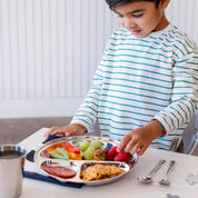 Child arranging fruits and snacks on a stainless steel divided plate with grapes, strawberries, peppers, tomoatoes, and pretzels.