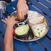 Children's hands reaching for food on a divided round stainless steel plate with various healthy foods and an 8 ounce bowl and 10 ounce cup. 