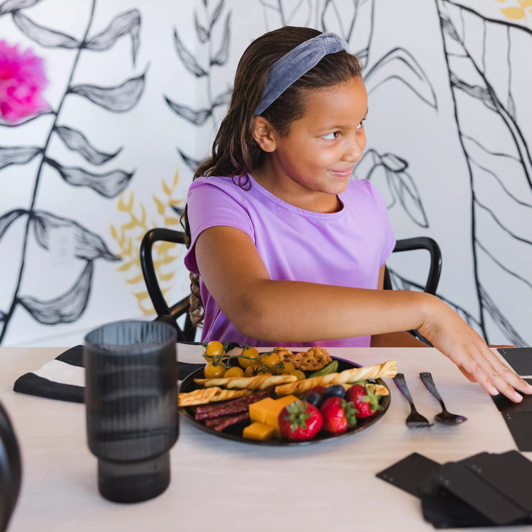 Young girl sitting at a table with a plate of food, wearing a purple shirt and a headband. Her plate in Ahimsa's graphite, stainless steel Purposeful Plate which is 9" as well as a fork and spoon - all dishwasher safe and made of stainless steel. The plate has strawberries, crackers, cheese, and tomatoes.