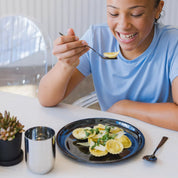 Teenager enjoying a meal of ravioli with a fork, sitting at a table with a plate of ravioli and a cup. The dishes are Ahimsa stainless steel dishes and dishwasher safe. The healthiest choice for families!