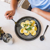 Teenager about to eat a stainless steel plate of pasta with a fork on a white tablecloth. The dishes are graphite or dark gray - they match your kitchen - plus kids love how they look and how shiny they are!