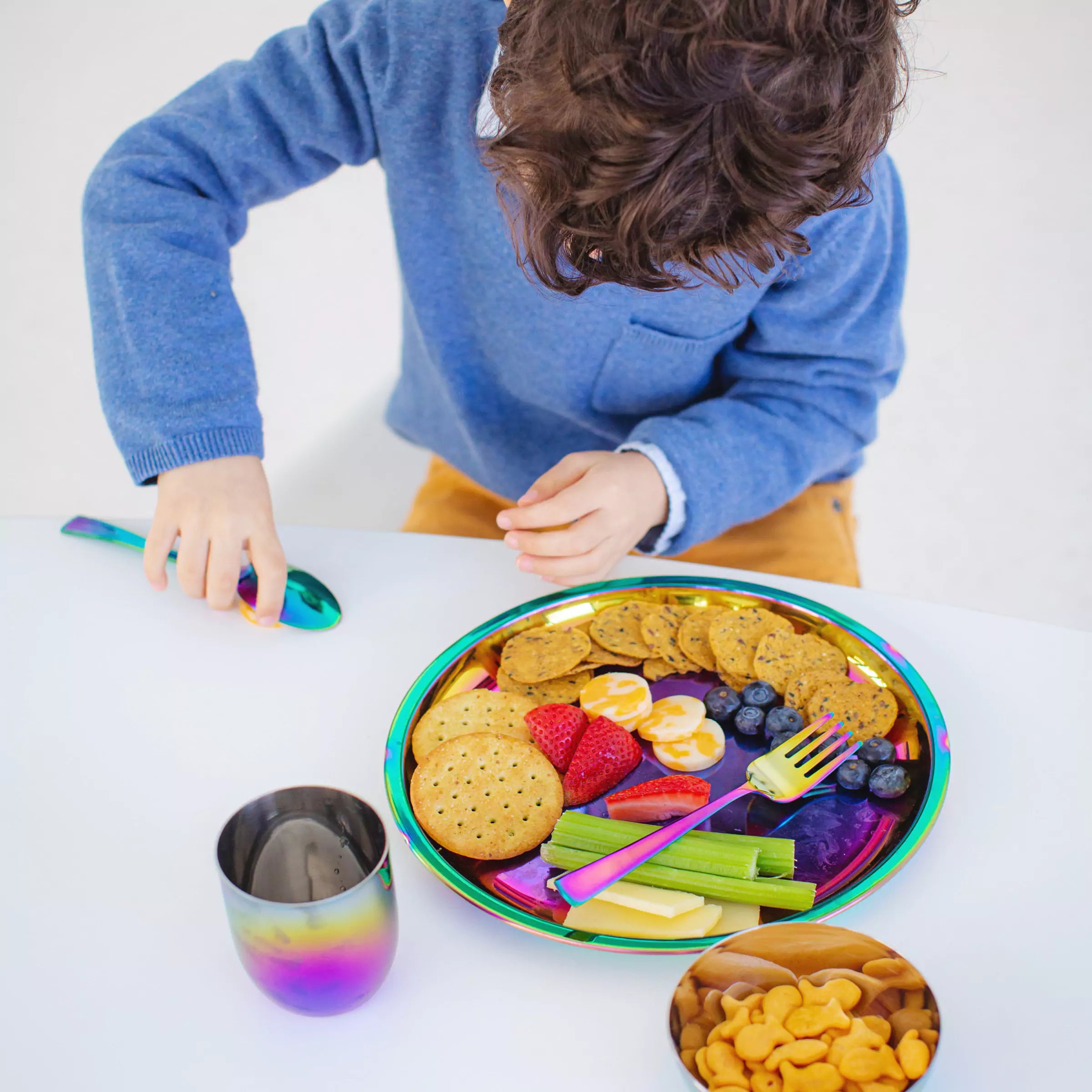 Little boy at a table with Ahimsa's Rainbow 9 inch stainless steel Purposeful Plate with food, 1 spoon, 1 fork, an 8 ounce cup and an 8 ounce bowl.