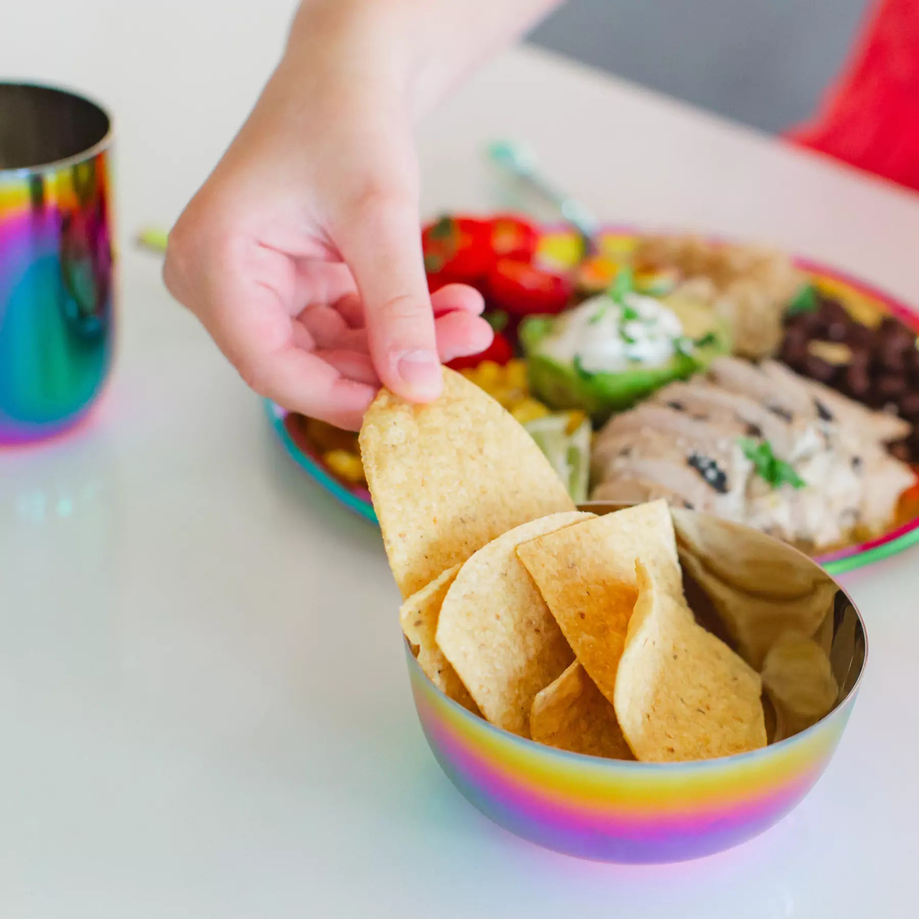 Child's hand reaching for a tortilla chip from a rainbow-colored stainless steel bowl with a colorful plate of food in the background.