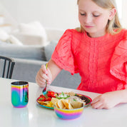 Child in a red dress eating a colorful meal at a table with a rainbow cup, plate and bowl. All Ahimsa dishes are dishwasher safe and free of harmful chemicals