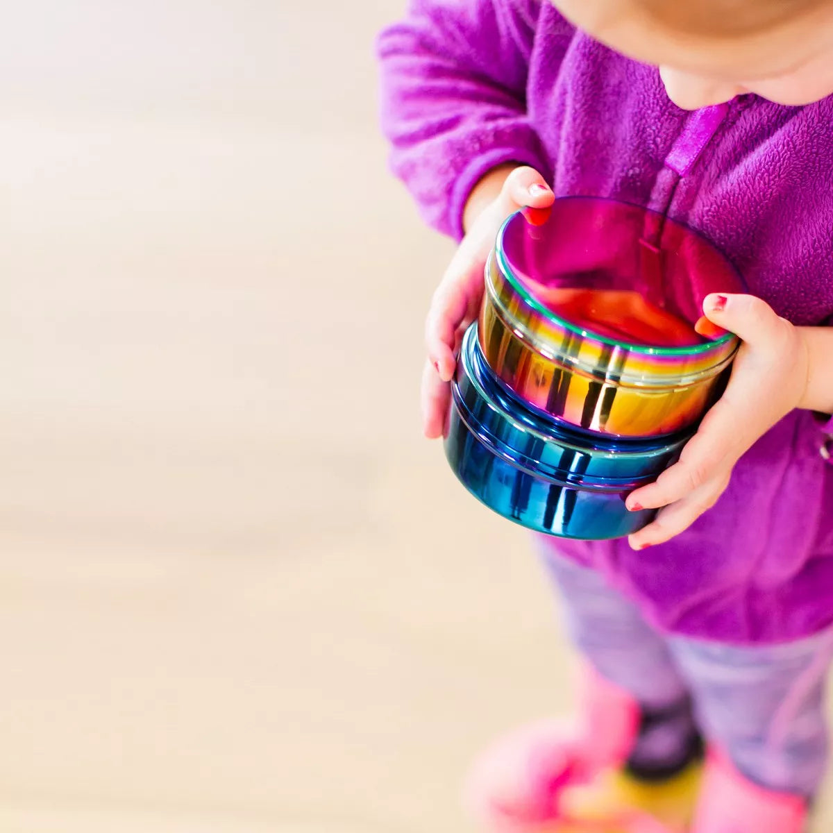 A child in a purple fleece sweater holds a stack of three iridescent rainbow-colored stainless steel food containers with a glossy finish - getting ready to pack her lunch.